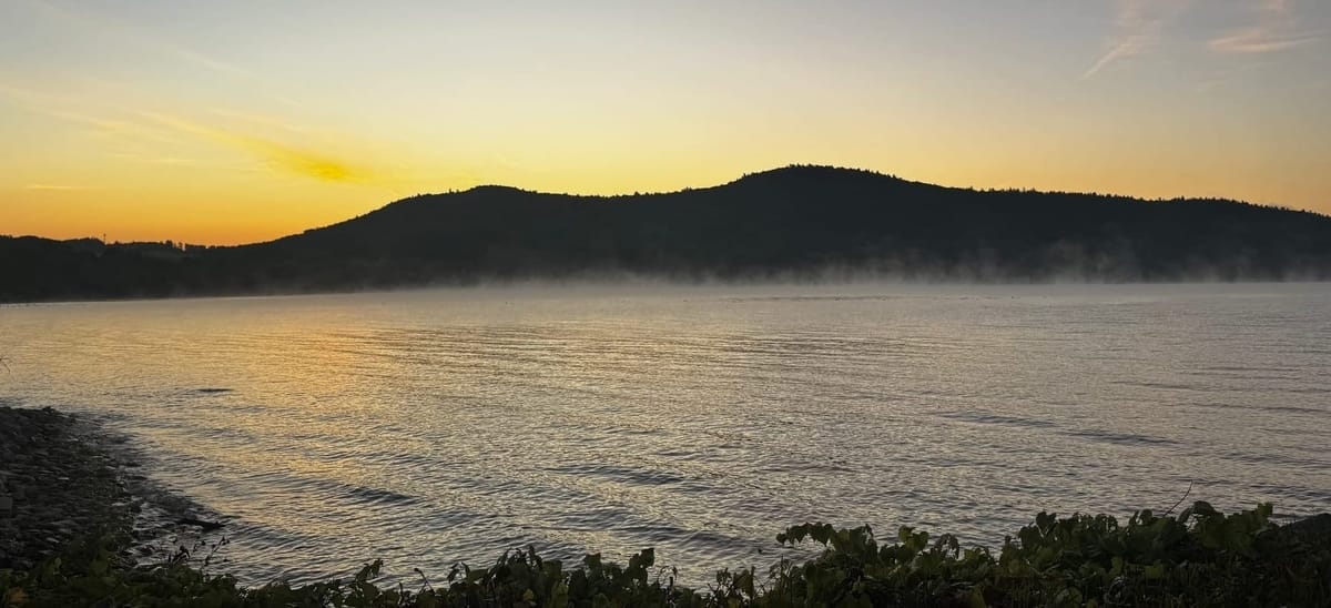 Sunrise over misty lake with silhouetted hills. Golden and orange sky reflected in calm water, morning fog hovering over surface, dark vegetation in foreground, wispy clouds above.